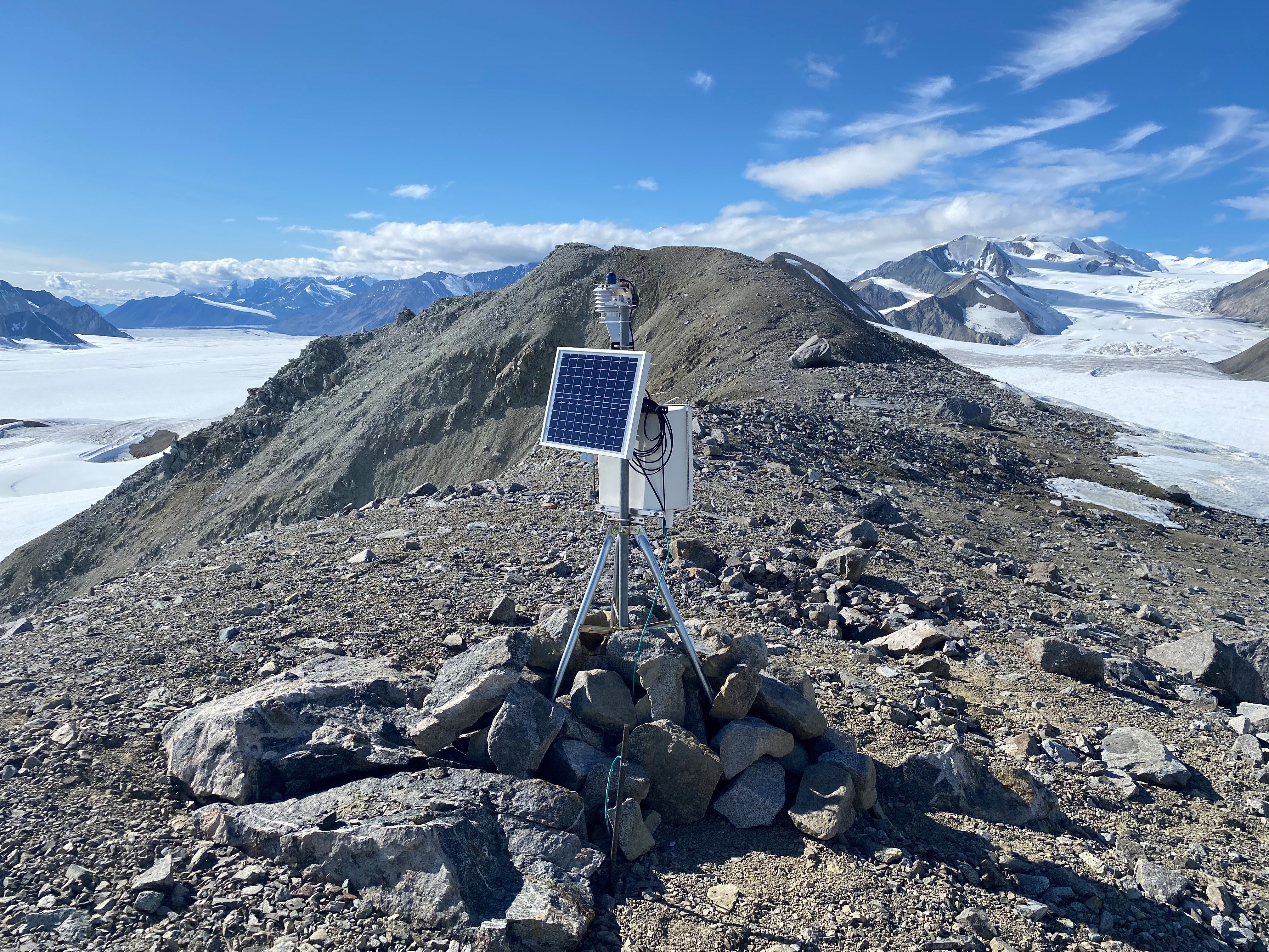 A pile of rocks holds a tripod mounted with a square solar panel on a metal box The view behind is a rocky, mountainous landscape.