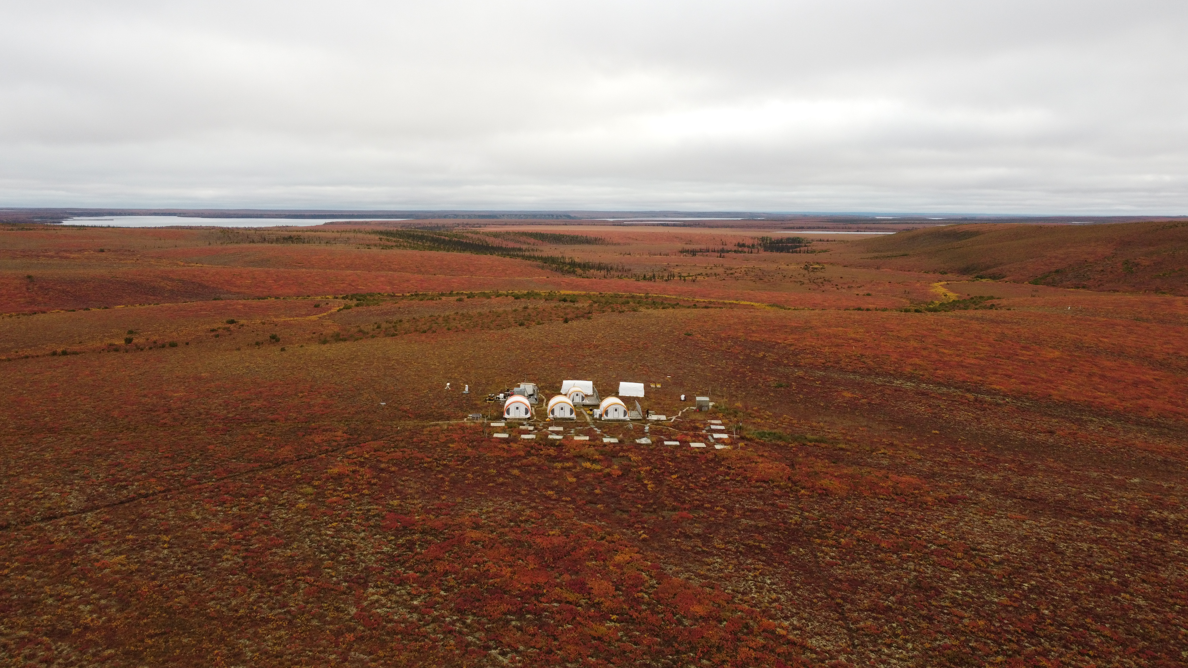 Aerial view of tundra in fall with a collection of six large buildings with smaller buildings in front. Lake in the far-right background.
