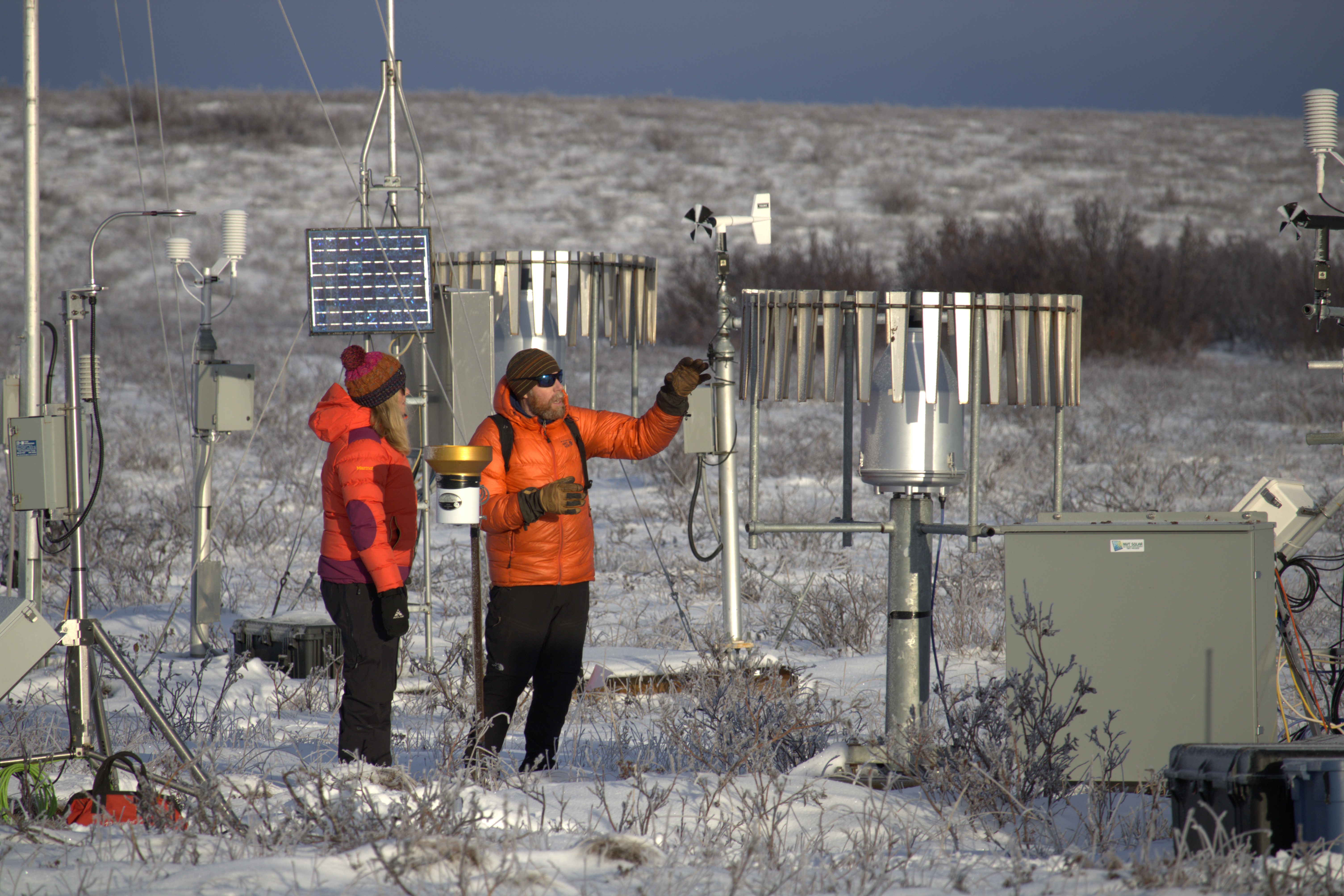 Two people, in winter parkas and gear, stand looking at tall, metal instruments in the snow-covered tundra.