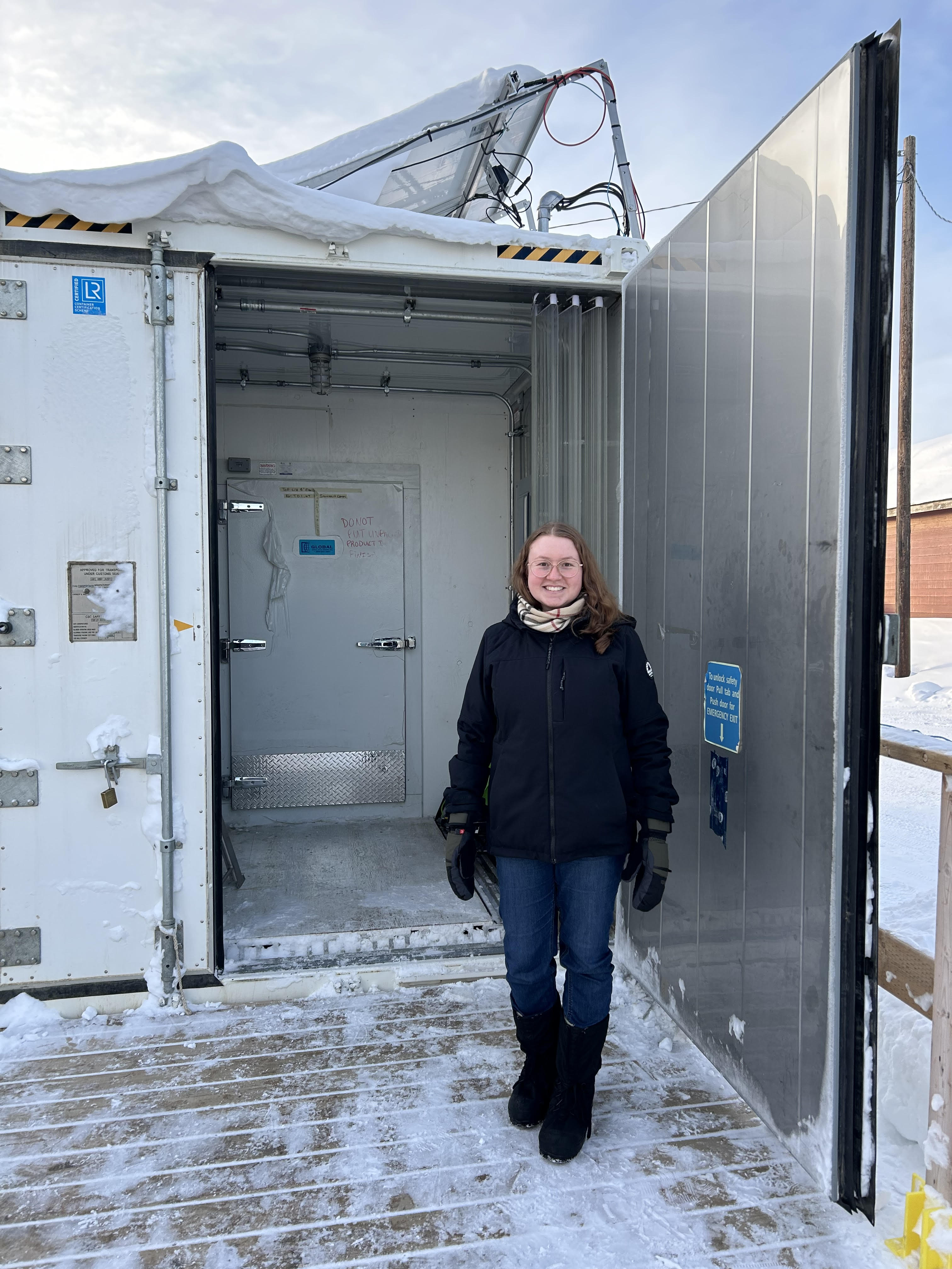 A woman stands outside beside the open door of a large metallic box.