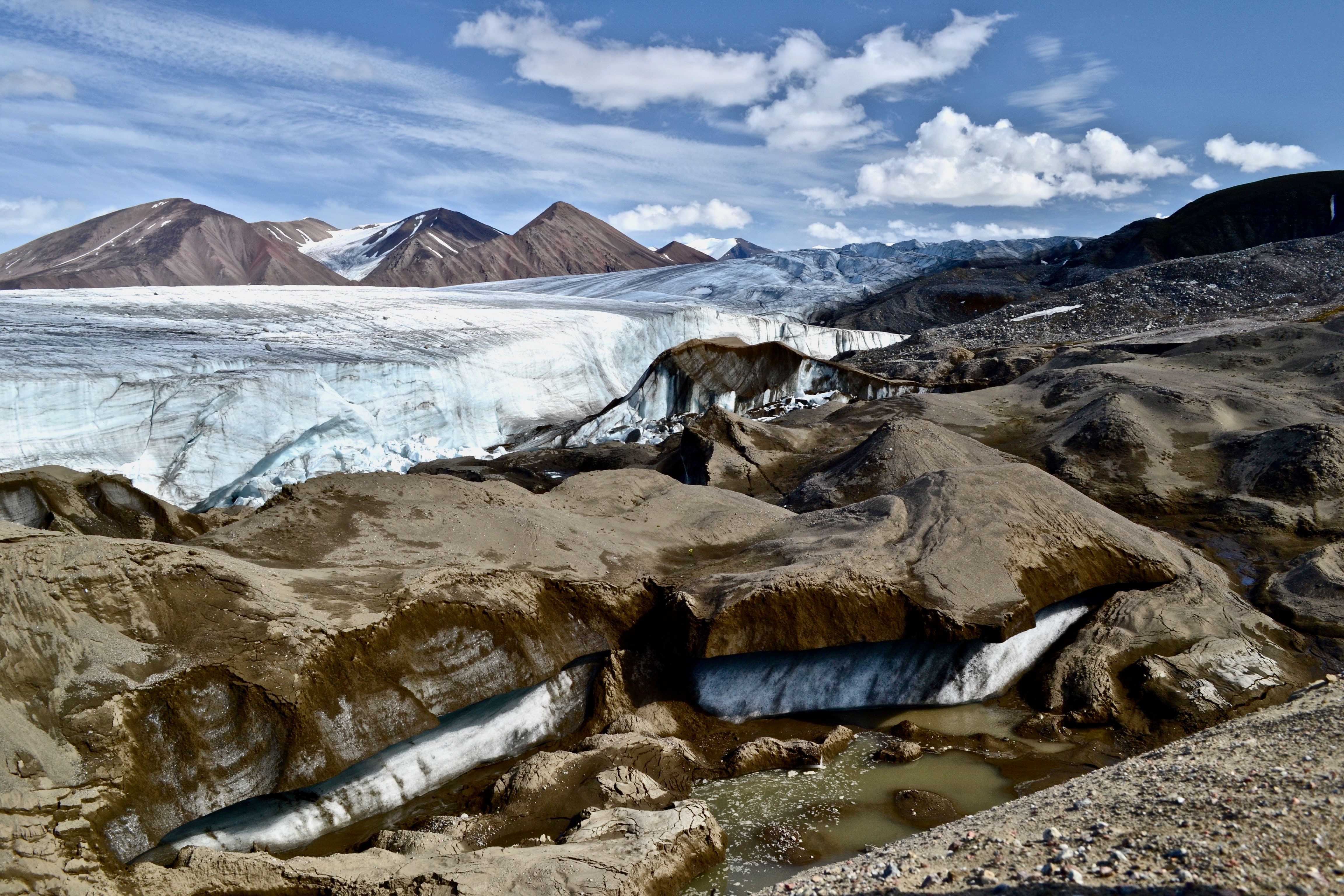 A scenic view with mountains in the background. A thick expanse of ice stretches across the middle of the photo with thick, dirt covered ice in the foreground.