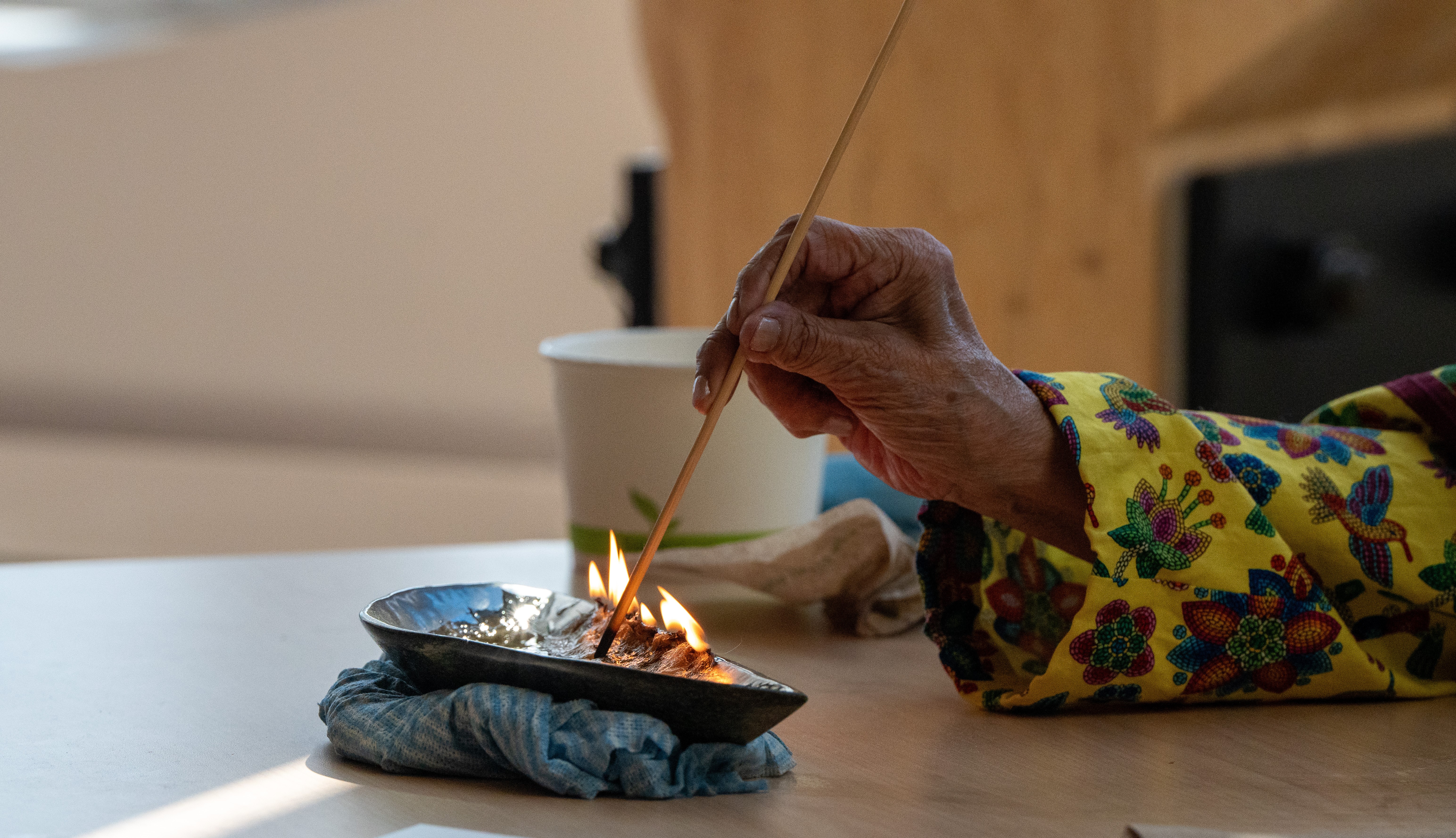 An old woman’s hand pokes a small line of fire in a shallow, stone dish with a long, thin stick.