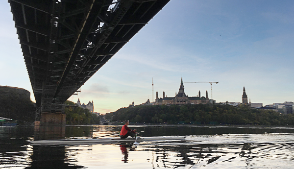 A view of a bridge from underneath with a rower on the river, and the Parliament Buildings in the background.