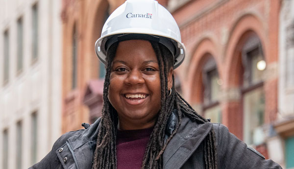 A woman in a construction helmet smiling and standing in front of a building.