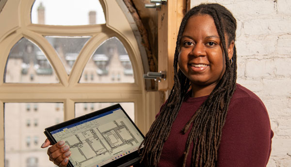 A smiling woman standing next to a heritage window and holding a computer with data on the screen.