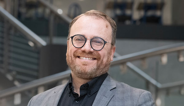 A man with glasses smiles as he stands in front of an interior staircase.
