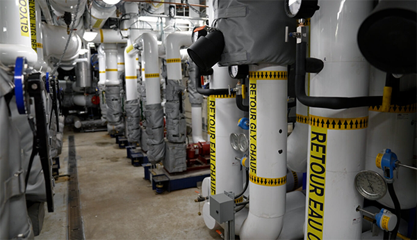  Une rangée de gros tuyaux de chauffage blancs avec une inscription noire sur fond jaune dans une salle mécanique.