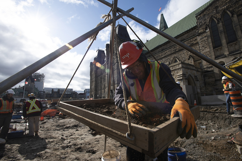 A person wearing a hard hat sorts through soil with the Centre Block in the background.