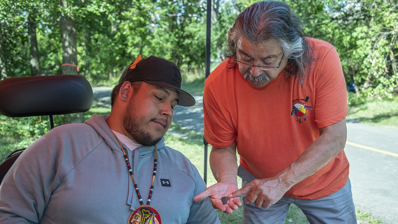 An older person shows a younger person a small stone sample.