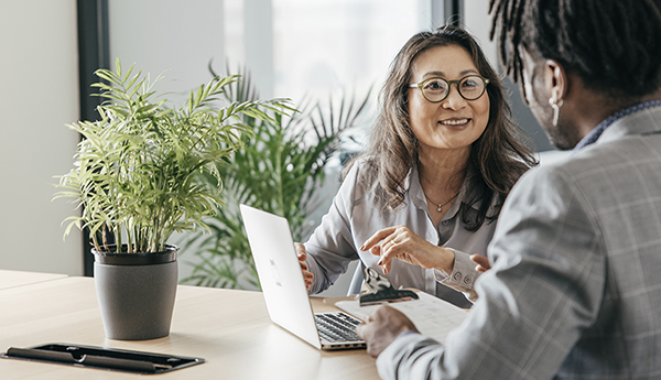 A photo of an Asian woman and a Black individual having a discussion at a table over an open laptop.