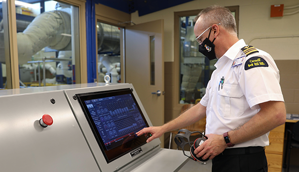 A person in a white naval uniform and face mask touches a computer monitor on a large console