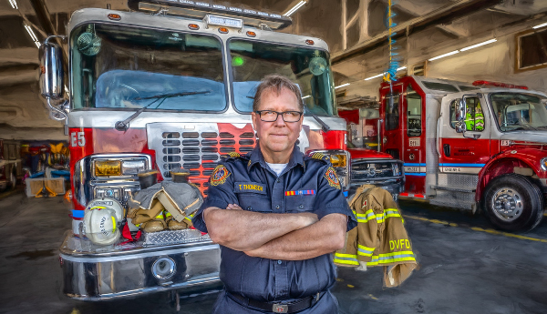 A firefighter in uniform stands with arms crossed inside a fire station, in front of a large fire truck.