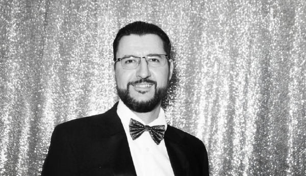 Black and white portrait of a bearded, smiling man wearing glasses and dressed in a tuxedo.
