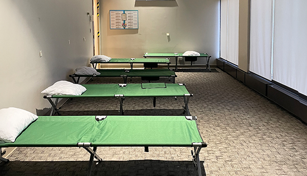 A row of green fabric cots in a small, carpeted room with windows.