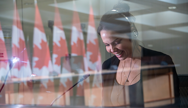 A smiling woman with a headset is seated in front of a microphone and behind a window reflecting a row of Canadian flags.