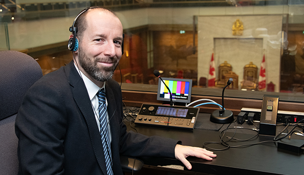 A smiling man with a headset sits in front of a microphone and screen in a glass office overlooking a room.