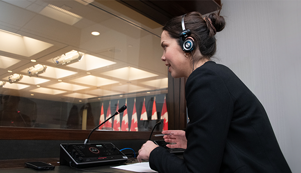 Profile of a focused woman with a headset, sitting in front of a microphone and screen in a glass office overlooking a room with a row of Canadian flags.