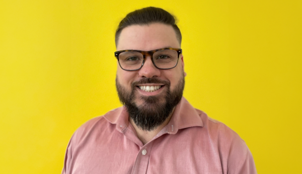Portrait of a smiling man with a beard, glasses and a pink shirt, on a yellow background.