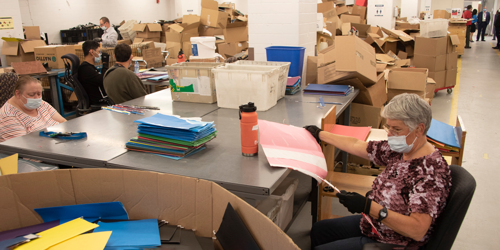 Masked employees sitting at a work station dismantling a paper file folder surrounded by piles of cardboard boxes.