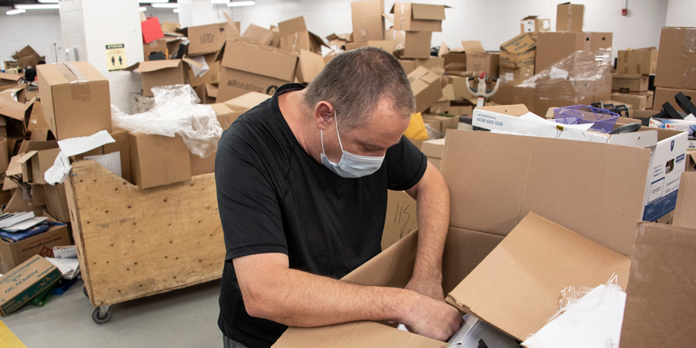 Masked person sorting materials in a box surrounded by piles of cardboard boxes.