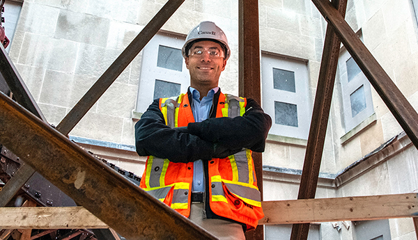 A young man in a construction vest and helmet standing with arms crossed among building beams.