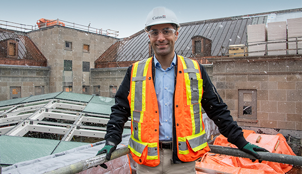 Un jeune homme vêtu d'un gilet de sécurité et d'un casque est debout sur le toit d’un édifice en chantier.