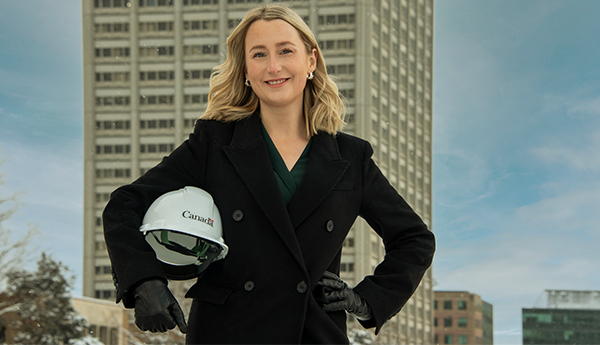 A young woman standing outside in front of a tall building and holding a construction helmet.