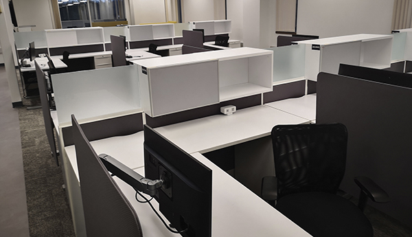 A room with rows of white desks with screens and a window in the back.