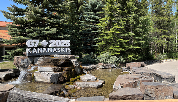 A G7 2025 Kananaskis sign by a waterfall and pond under a clear sky, surrounded by rocks and evergreens.