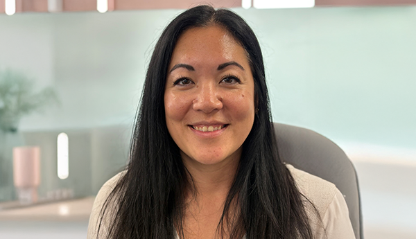 A woman with long dark hair sitting indoors against a light-coloured wall with rectangular decorations.