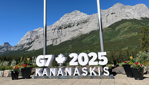 A G7 2025 Kananaskis sign in front of a mountain landscape under a clear blue sky, with flower pots in the foreground.
