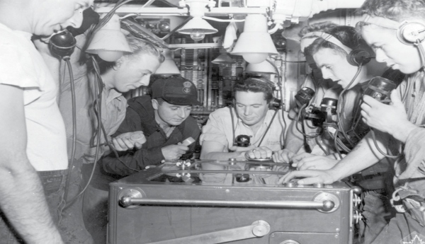 A black and white photo showing a group of men in uniform and wearing earphones huddled over a table on a ship.