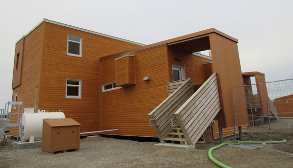 Square wooden building with a staircase in front and a white tank on the left