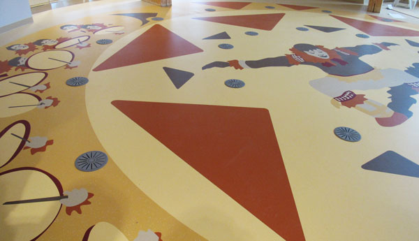 Inuit artwork on the floor showing orange triangles of various sizes and an Inuit in traditional costume in the middle, surrounded by faces and drums