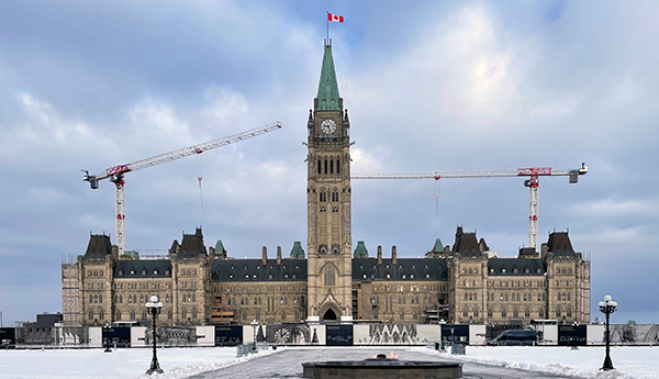 Front view of Parliament Hill in winter with the Canadian flag flying on the Peace Tower and cranes on either side of the tower.