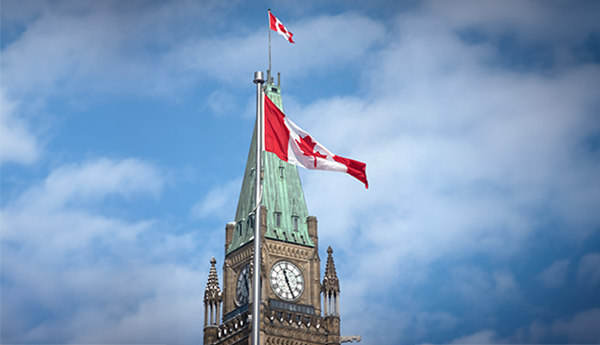 A Canadian flag raised on a pole, with a different Canadian flag flying on the Peace Tower behind the pole in the background.
