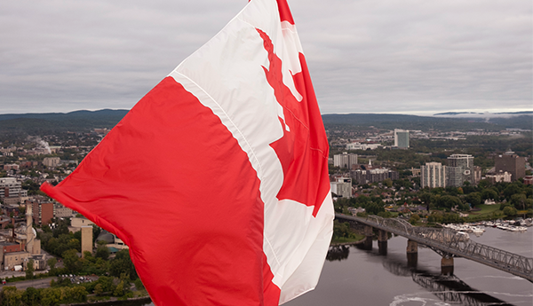 Close-up view of the Canadian flag flying on Parliament Hill. A bridge, a river and a town are in the background.