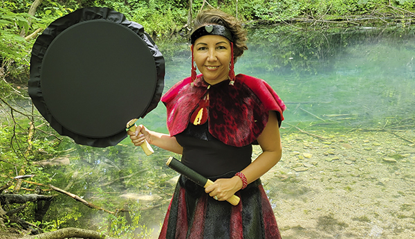 Vanessa Brousseau debout devant un cours d’eau vêtue d’une robe rouge et noir en fourrure et tenant un tambour noir.