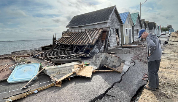 A man examines a cracked pavement and debris next to houses along a body of water.