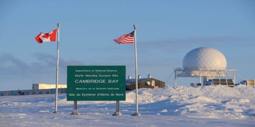 A snowy landscape with radar facilities in the background, and Canadian and American flags in the foreground, together with a sign saying Cambridge Bay