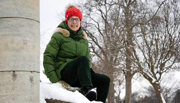 Cathryn Bjerkelund wearing a green winter coat and a red hat is sitting next to a concrete column