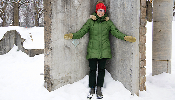 Cathryn Bjerkelund wearing a green winter coat and a red hat is standing in the snow against a concrete wall