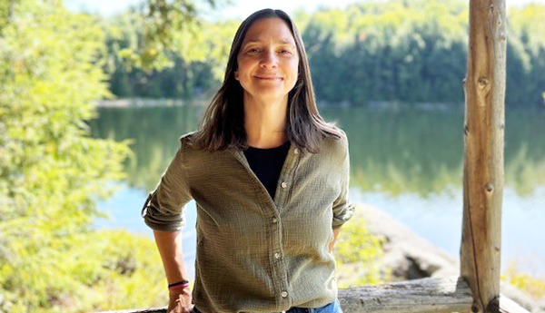 Smiling woman with long hair, standing in front of a lake and trees.