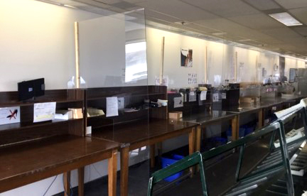 Suite of tables along a wall, divided by plexiglass. There is office supplies on the tables.