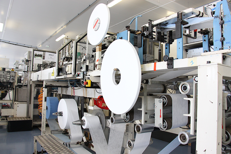 A large automated machine with a series of large bobbin-like rollers containing material to be made into personal protective equipment.