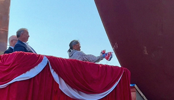 Une femme fracasse une bouteille de champagne contre la coque d'un grand navire rouge tandis que 2 hommes regardent derrière elle.
