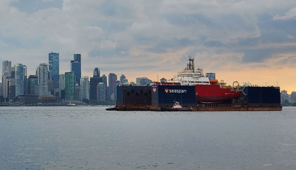 Un navire rouge et blanc sur une barge naviguant sur une grande étendue d'eau avec la ligne d'horizon d'une ville en arrière-plan.