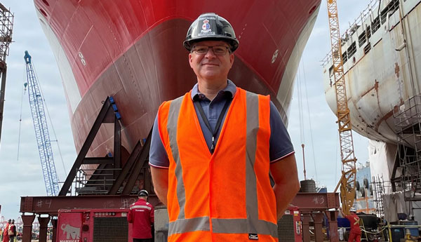 Un homme portant un casque de protection et un gilet de sécurité se tient devant un grand navire rouge.