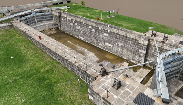 A 3D illustration of a fluvial canal made of grey stone with 2 metal doors surrounded by grass.