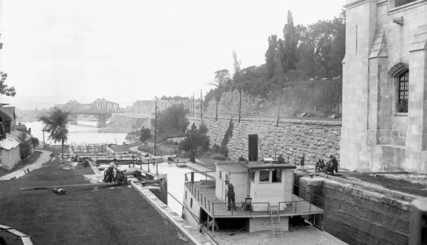 A black and white photograph of a fluvial canal with a boat crossing, some workers on the ground, as well as a metallic bridge further in the background.